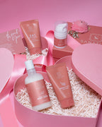 A collection of sanatlyn skincare products including a body wash, face cleansers, and a cream, all packaged in pink containers and presented in a heart-shaped box with 'thank you' notes.