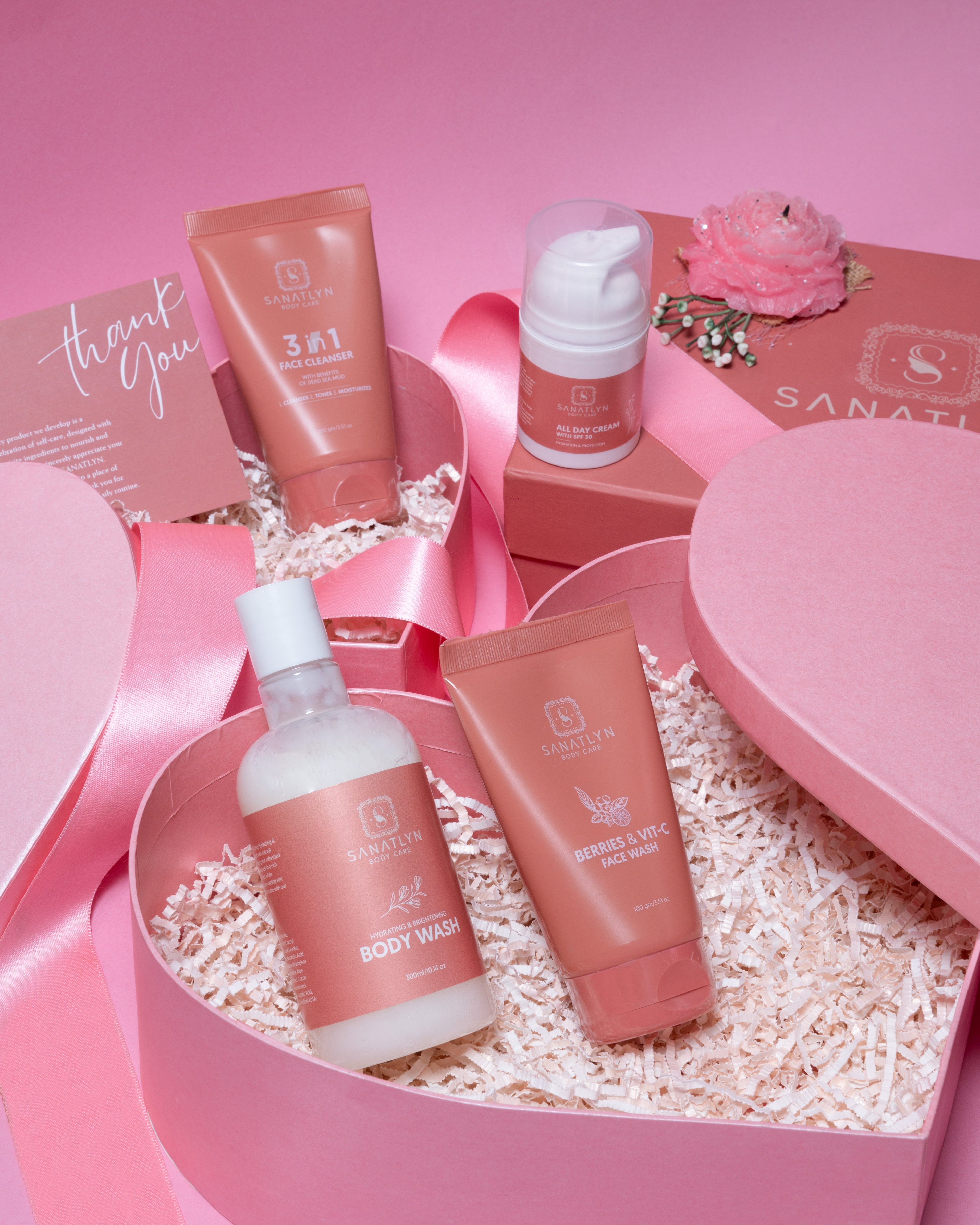 A collection of sanatlyn skincare products including a body wash, face cleansers, and a cream, all packaged in pink containers and presented in a heart-shaped box with 'thank you' notes.